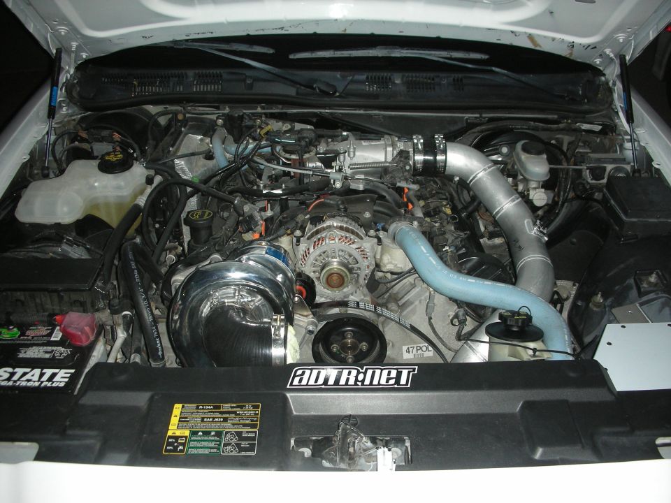 Engine bay with a chrome supercharger, silver intake piping, and a black radiator cover with text