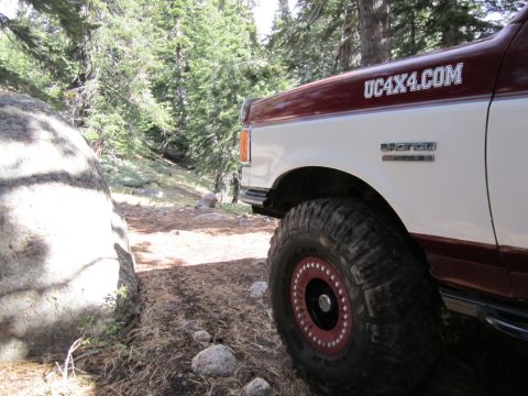 1991 Ford Bronco - A 1991 Ford Bronco is parked off-road, with its front fender and large tire visible against a backdrop of trees and rocks.