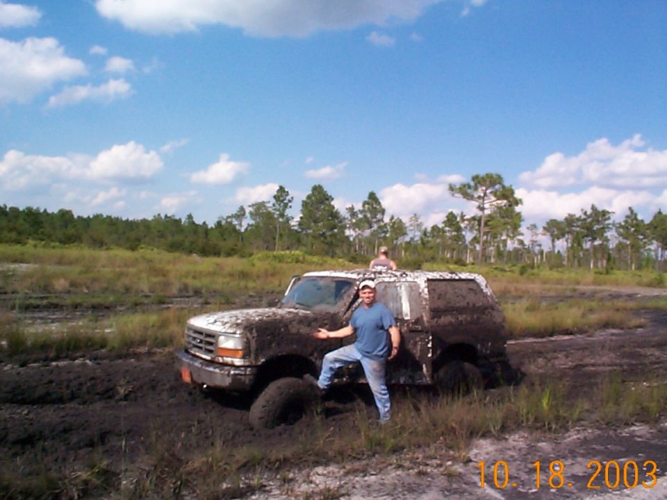 Man in jeans stands beside a mud-covered full-size Bronco stuck in a muddy field