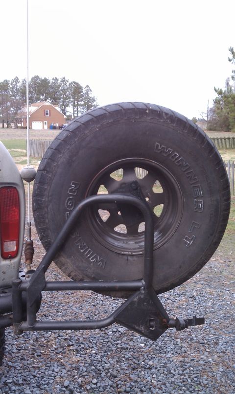 1979 Ford Bronco - The rear of a vehicle shows a spare tire mounted on a custom carrier, with a house visible in the distance.