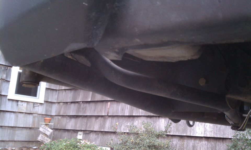 Underside view of a vehicle's frame and exhaust pipes, with a house in the background