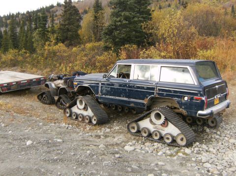 2000 Ford E350 - A modified Jeep Grand Wagoneer with tank treads is parked on a gravel surface, with a trailer attached.