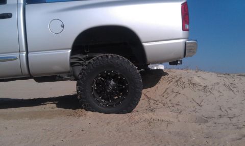 2005 Dodge 1500 - Rear view of a silver pickup truck with large off-road tires, parked on a sandy dune under a clear blue sky.