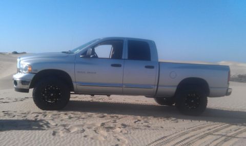 2005 Dodge 1500 - Silver pickup truck with black rims parked on sand dunes under a clear blue sky.