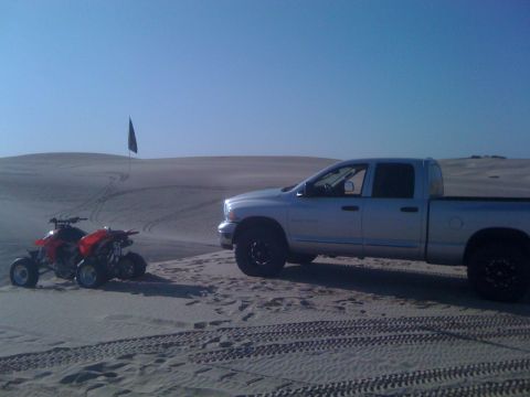 2005 Dodge 1500 - A silver pickup truck and two red ATVs are parked on sand dunes under a clear blue sky.