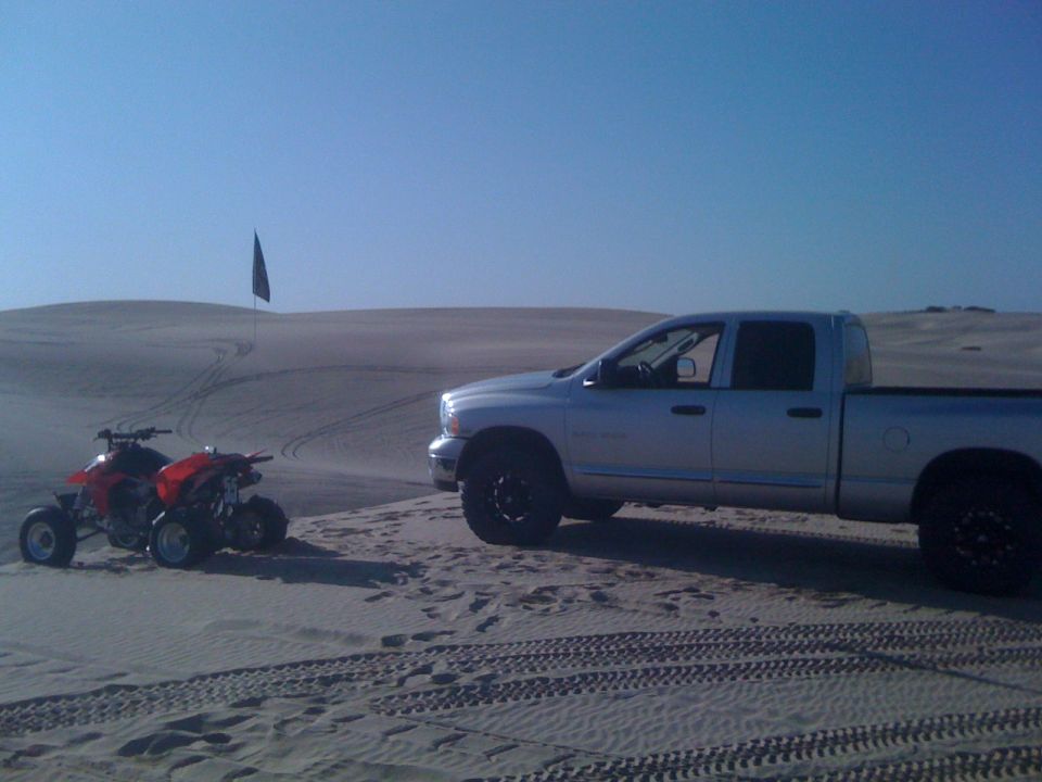 Silver pickup truck parked on sand dunes next to two red ATVs