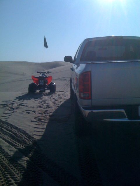 2005 Dodge 1500 - A silver 2005 Dodge 1500 pickup truck is parked on sand dunes, with a red ATV in the background under a clear blue sky.