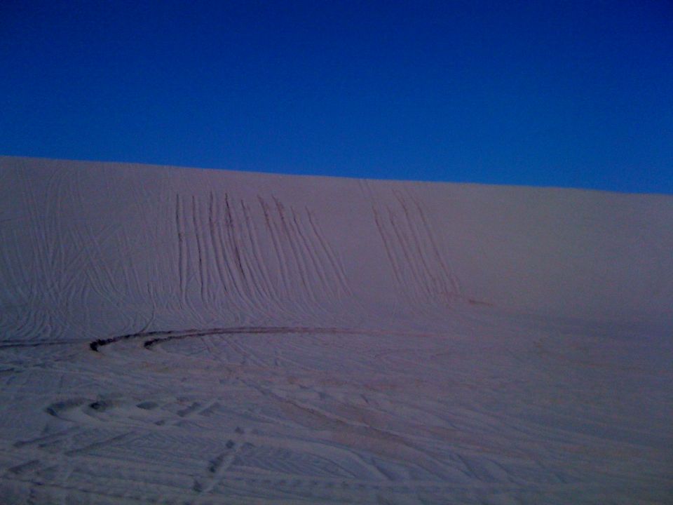 Sand dunes with tire tracks under a clear blue sky. Supermotors.net logo in lower right