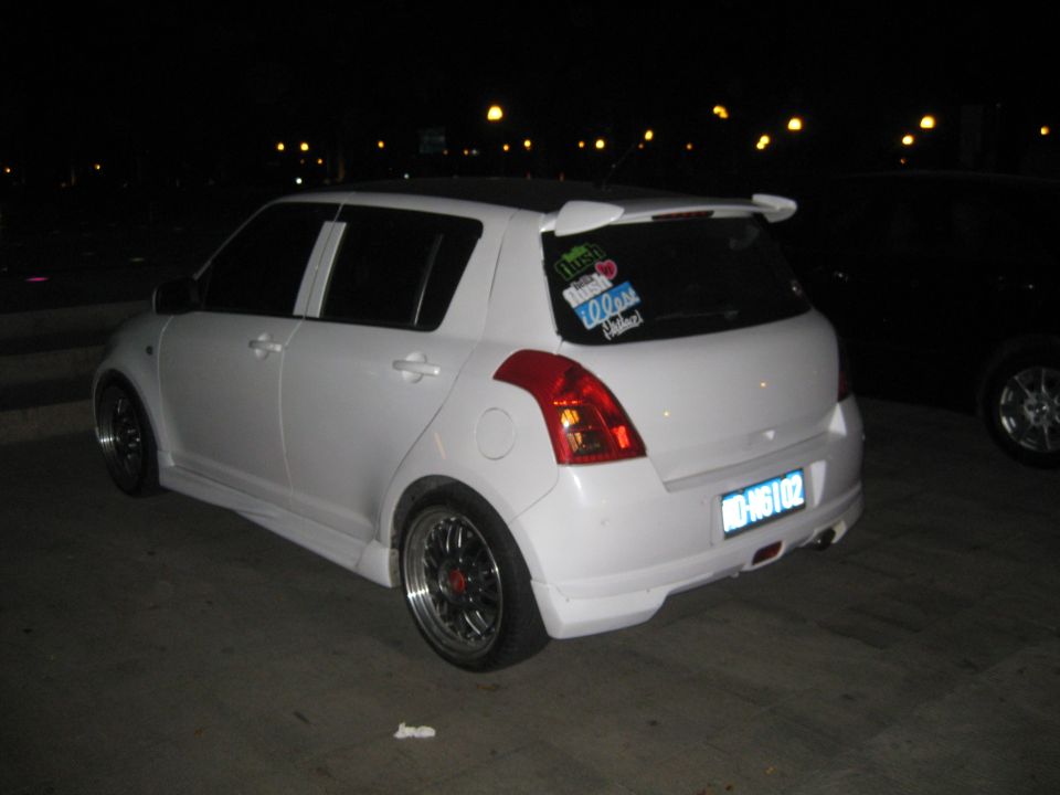White hatchback with aftermarket spoiler and wheels parked at night. Stickers on rear window