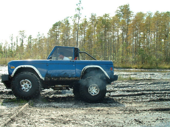 Blue topless early Ford Bronco on tall mud tires driving through a muddy pit