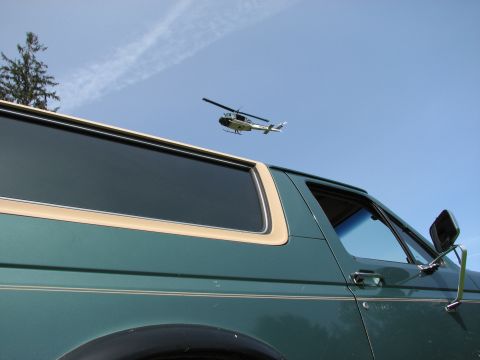 1990 Ford Bronco - A helicopter flies over a green SUV, seen from a low angle against a blue sky.