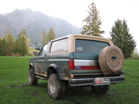 1990 Ford Bronco - A green and tan 1990 Ford Bronco is parked in a grassy field with mountains and a helicopter in the background.