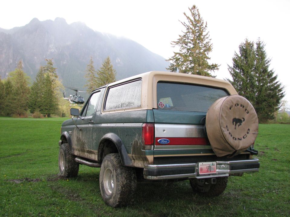 Rear view of a green and tan 1990 Ford Bronco with a spare tire cover and a helicopter in the background