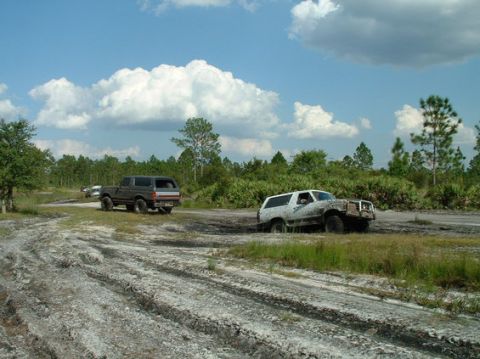 1992 Ford Bronco - Two Ford Broncos navigate a muddy off-road course under a partly cloudy sky.