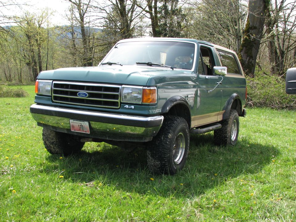 Green Ford Bronco with tan stripe, lifted on off-road tires, parked in a grassy field