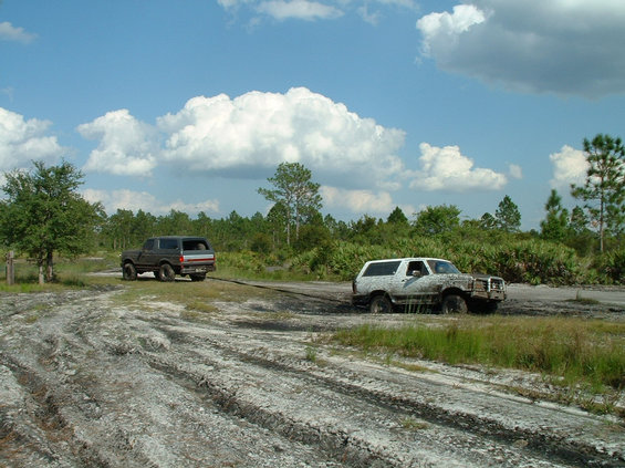 Mud-covered white Bronco stuck in a mud pit being strapped to a dark Bronco on a sand trail