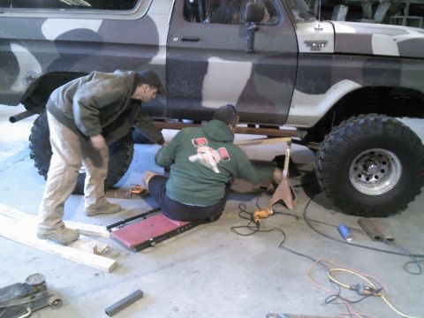 1979 Ford Bronco - Two individuals work on the undercarriage of a 1979 Ford Bronco, which features a distinctive camouflage paint scheme.