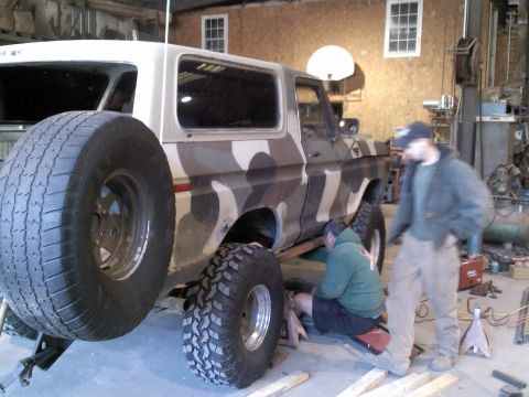 1979 Ford Bronco - Two mechanics work on a Ford Bronco in a garage, which has camouflage paint and a rear-mounted spare tire.