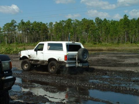 1992 Ford Bronco - A white 1992 Ford Bronco navigates a muddy terrain with a spare tire on the back.