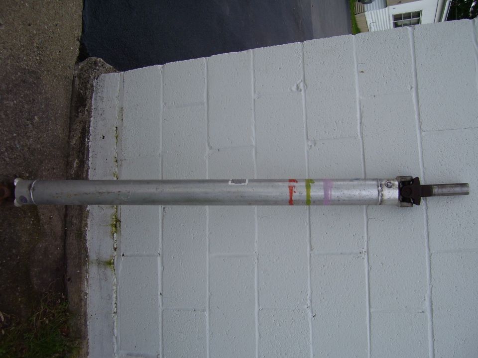 Silver one-piece driveshaft with U-joint leaning against a white cinder block wall