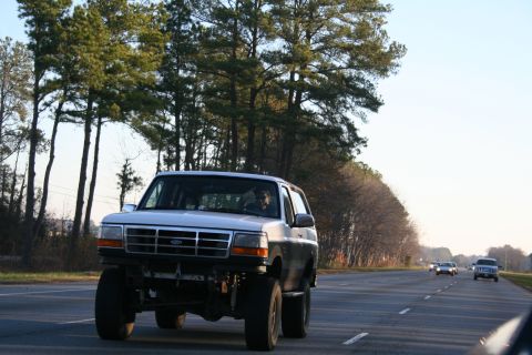 1982 Ford Bronco - Lifted Ford Bronco driving on a highway with trees lining the road.