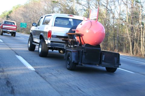 1982 Ford Bronco - Silver Ford Bronco tows a trailer with a large pink smoker down the highway, with another truck visible ahead.