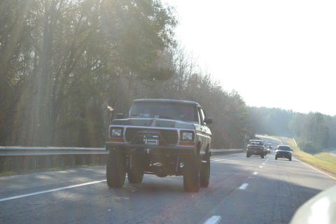1982 Ford Bronco - A lifted Ford Bronco drives down a highway, with other vehicles in the distance.