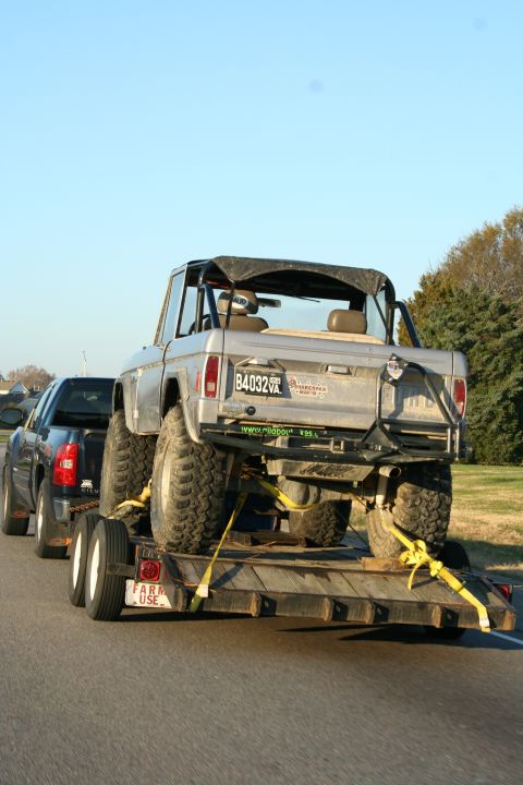 1982 Ford Bronco - A silver convertible SUV is towed on a trailer behind a dark pickup truck.