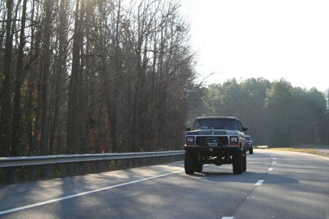 1982 Ford Bronco - A dark SUV with a lifted suspension drives down a road, with another vehicle visible in the distance.