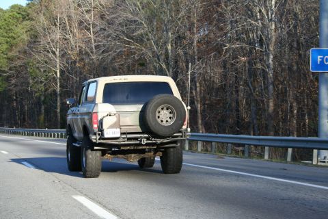 1982 Ford Bronco - Rear view of a lifted 1980s Ford Bronco with a spare tire on the back, traveling on a road.
