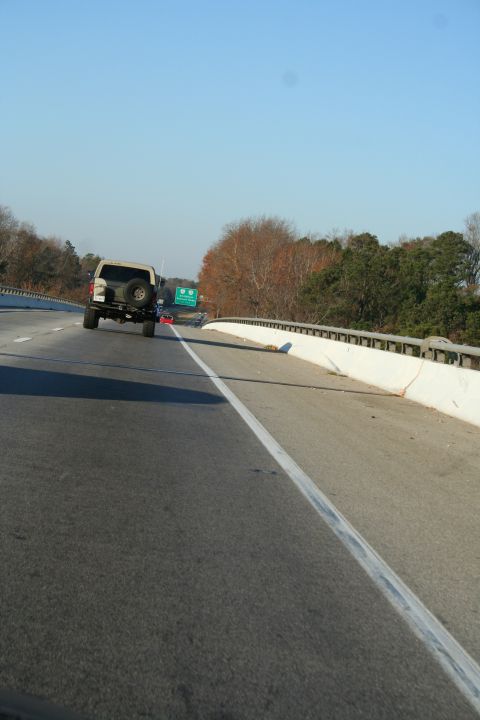 1982 Ford Bronco - A tan SUV with a spare tire mounted on its rear is seen driving on a highway under a clear blue sky.