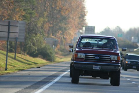 1982 Ford Bronco - A red pickup truck drives down the road with trees and other vehicles in the background.