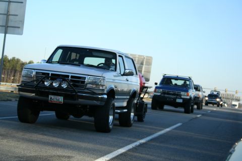 1982 Ford Bronco - A silver SUV, likely a 1992-1996 Ford Bronco, leads a procession of vehicles on a highway, towing a trailer.
