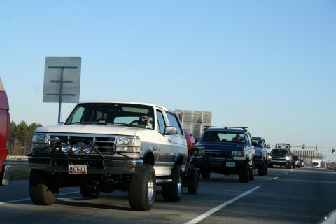 1982 Ford Bronco - A line of vehicles, including a lifted white Ford Bronco, travels down a highway under a clear blue sky.