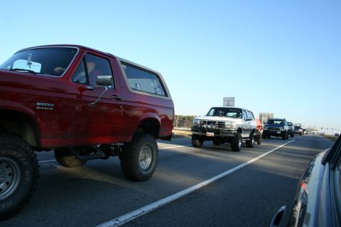 1982 Ford Bronco - A convoy of SUVs, including a red Ford Bronco, travels down a highway under a clear blue sky.