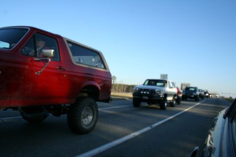 1982 Ford Bronco - A red 1982 Ford Bronco leads a line of trucks on a highway under a clear blue sky.