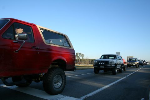 1982 Ford Bronco - A convoy of trucks, including a red Ford Bronco, travels down a highway under a clear blue sky.