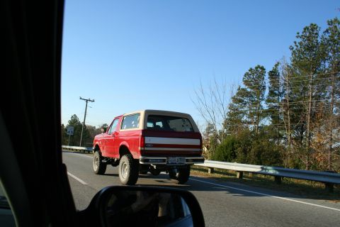 1982 Ford Bronco - A red Ford Bronco with a tan top is driving down a highway on a clear day.
