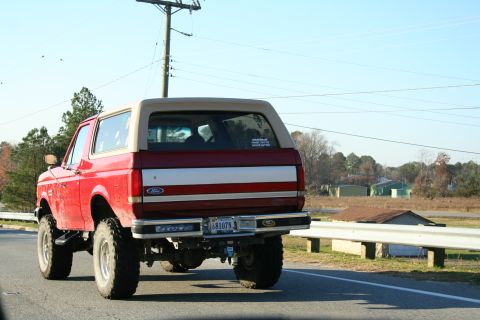 1982 Ford Bronco - This red Ford Bronco with a tan top is seen from the rear, driving on a road with a lifted suspension.