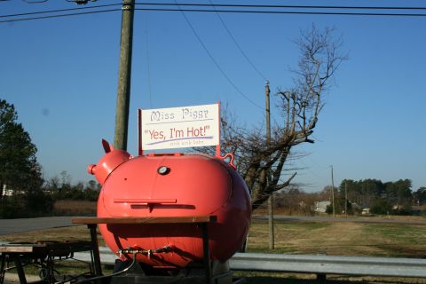 1982 Ford Bronco - A large, red, pig-shaped smoker sits outdoors under a clear blue sky, displaying a sign that reads 'Miss Piggy "Yes, I'm Hot!"'.