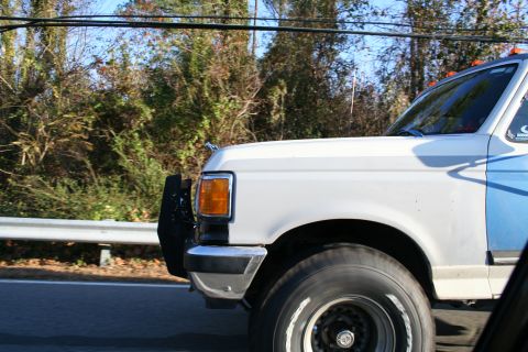 1982 Ford Bronco - A white and blue truck drives down the road, its side profile visible against a backdrop of trees.