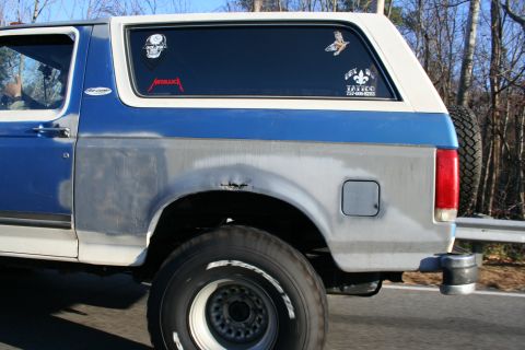 1982 Ford Bronco - A blue and white Ford Bronco is seen from the side, showing primer-gray bodywork and various stickers on the rear window.