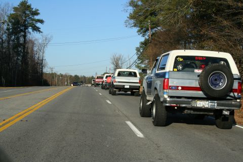 1982 Ford Bronco - A line of SUVs, including a lifted blue and silver model with a rear-mounted spare tire, travel down a two-lane highway.