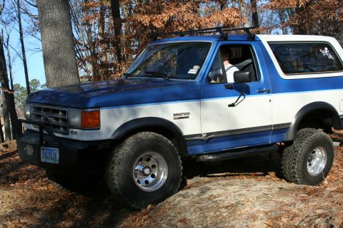 1982 Ford Bronco - This blue and white Ford Bronco, likely from the late 1980s, is equipped for off-road adventures with its large tires and roof rack.