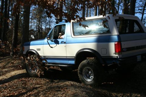 1982 Ford Bronco - A two-tone blue and white Ford Bronco is seen from the side on a wooded trail.