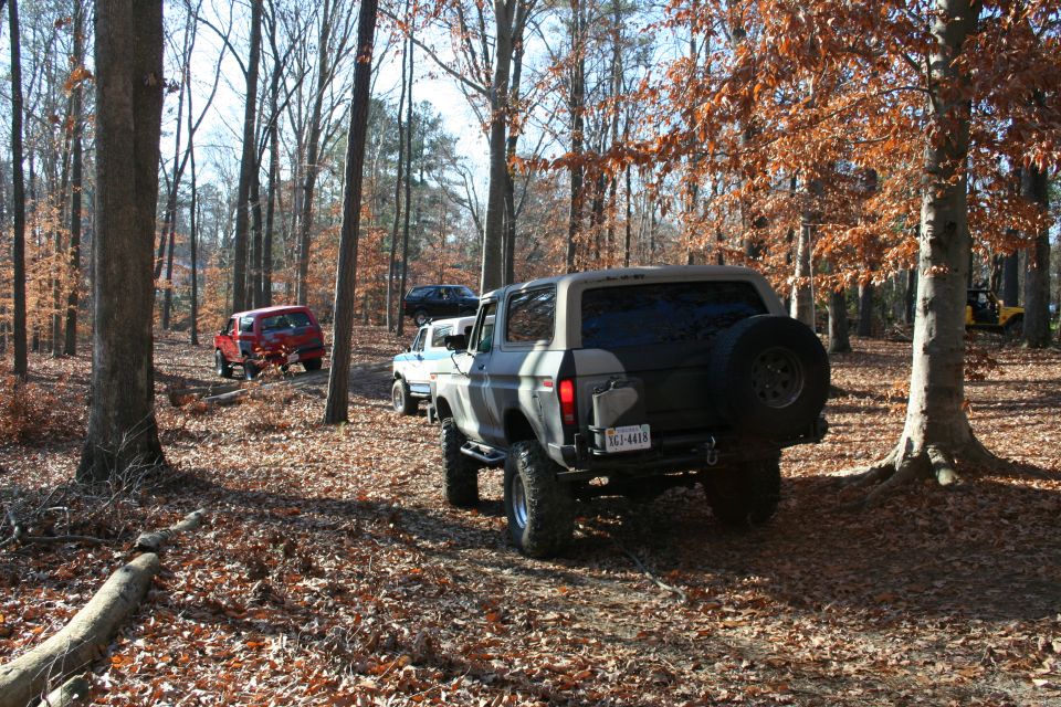 Gray Ford Bronco with spare tire on back, driving on a dirt trail in woods. Red SUV and white truck visible behind it
