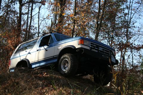 1982 Ford Bronco - A two-tone blue and white Ford Bronco navigates an off-road incline amidst autumn trees.