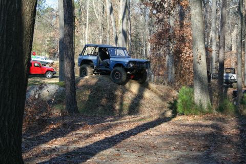 1982 Ford Bronco - Several off-road vehicles are parked in a wooded area, with one blue SUV climbing a dirt mound.
