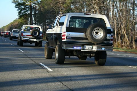 1982 Ford Bronco - A line of Ford Bronco SUVs travel down the road, one with a spare tire mounted on the back.