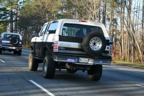 1982 Ford Bronco - Two Ford Broncos are seen driving on a road, with the lead vehicle partially obscured by the one behind it.